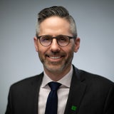 head and shoulders photo of a bespectacled man in a suit wearing a suit and tie smiles 